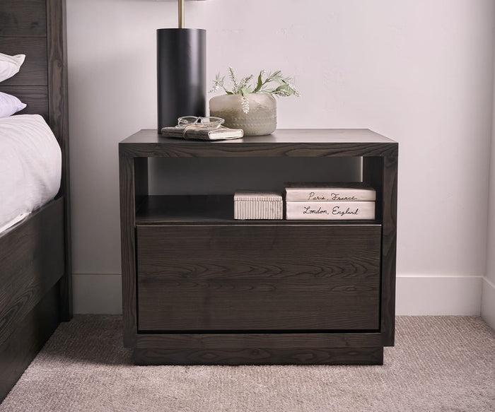 A dark wood nightstand with an open shelf holding books and a lower drawer sits beside a bed on a carpeted floor. On top, decor items include glasses, the Tula Marble Box With Lid, a book, a vase with greenery, and a tall black lamp. The handcrafted nightstand completes the cozy bedside setup.