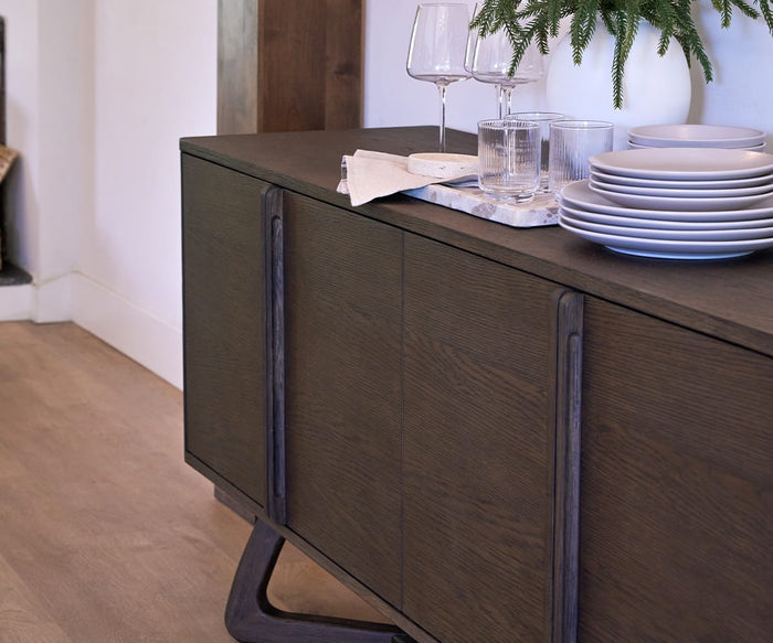 A dark wood sideboard holds stacked plates, glasses, wine glasses, and a white vase with greenery, beautifully accented by the Tula Rectangle Marble Tray displaying handcrafted serveware. The scene is set on a light wood floor in a modern dining area.
