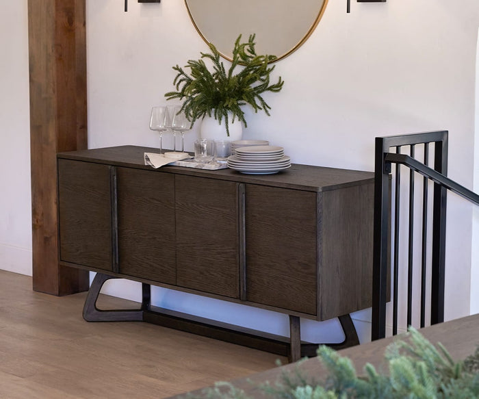 A dark wood sideboard with dishes, glasses, and the Tula Rectangle Marble Tray featuring handcrafted serveware sits against a white wall beneath a round mirror. A staircase railing is on the right and a wooden column on the left.