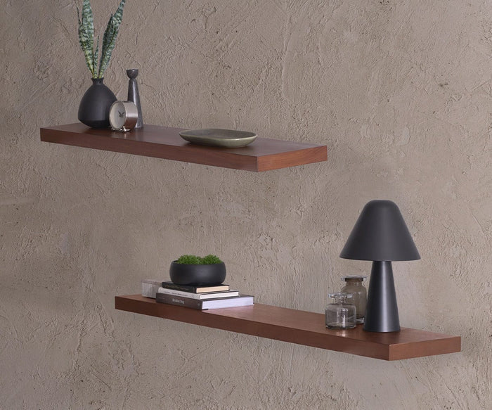 Two Ulin 24″ wooden floating shelves are mounted on a textured wall, displaying decorative items such as a lamp, small plants, books, a clock, vases, and a bowl. This multifunctional arrangement of Ulin 24″ Shelves enhances the minimalist and modern decor of the space.