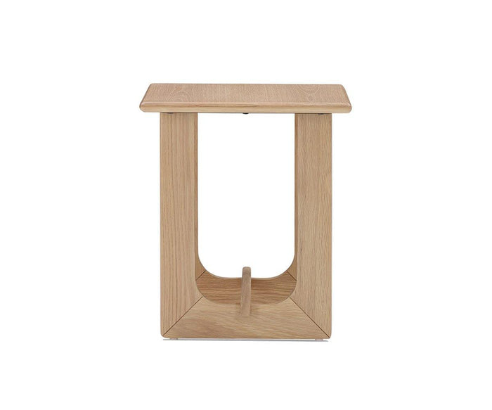 The Unni Side Table features a modern, light wood design with a square top and an open rectangular frame, highlighted by a curved base support that elegantly references classic mid-century style.