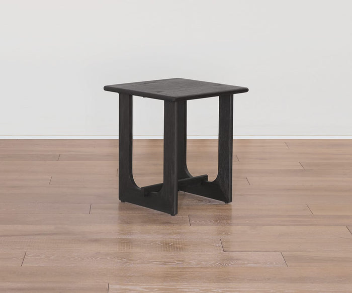 The Unni Side Table, featuring a compact black wooden design with a square top and open U-shaped legs, is shown on a light brown wood floor against a plain white wall.