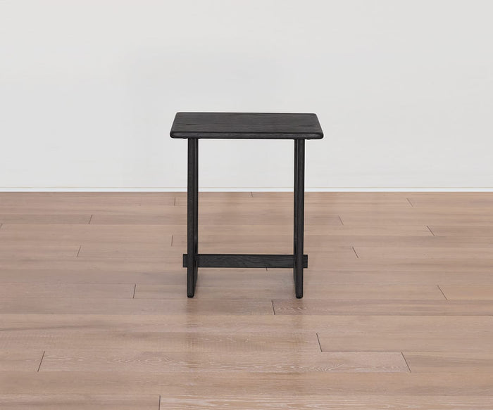A small, black Unni Side Table with a rectangular mid-century design and straight legs is placed on a light brown wooden floor against a plain white wall.