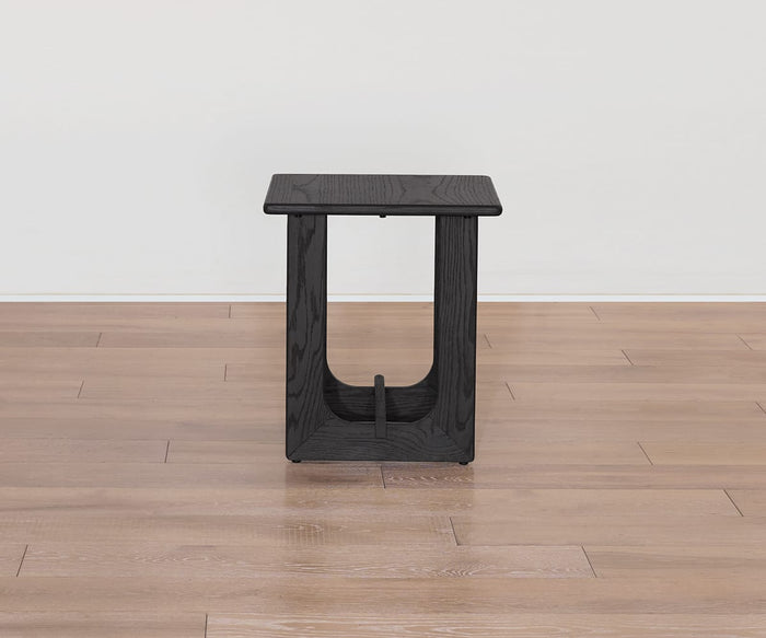 The Unni Side Table, a small and modern black wooden piece with an open base, stands on a light brown wooden floor against a plain white wall.