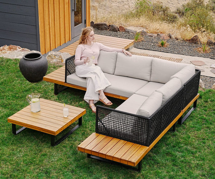 A woman sits on a modern outdoor sofa with light cushions, holding a drink. Beside her, the Vila Outdoor Square Coffee Table with a teak finish holds a pitcher and glass on the grassy yard next to a contemporary house.