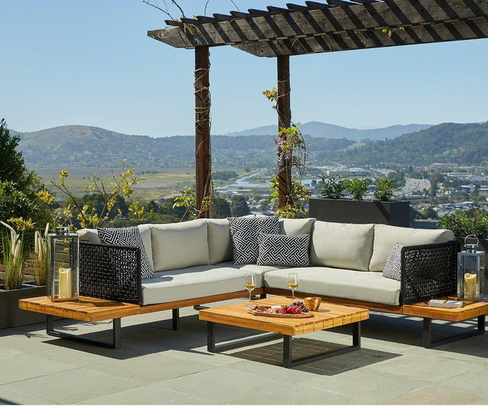 Outdoor patio with a modern sectional sofa and patterned cushions, featuring the Vila Outdoor Square Coffee Table crafted from acacia wood with a sleek teak finish, topped with drinks and snacks. Pergola overhead, scenic hills and a highway visible in the background.