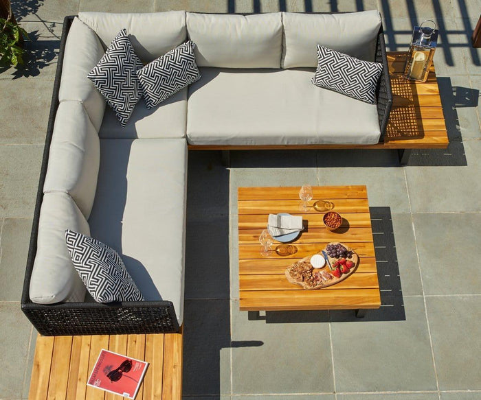 A patio featuring a gray cushioned L-shaped sofa with patterned pillows, the Vila Outdoor Square Coffee Table topped with snacks and wine, and a magazine resting on a side table.