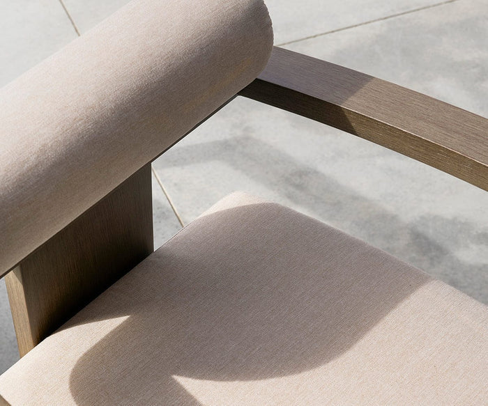 A close-up of the Vinna Outdoor Dining Chair showcases its light beige, weather-resistant fabric cushion and wooden armrest, casting a shadow on the tiled floor under bright sunlight.