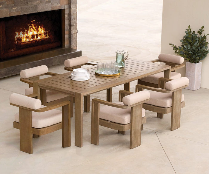 The Vinna Outdoor Dining Table, paired with six cushioned chairs, is arranged with plates, glasses, and a pitcher near a glowing fireplace and a potted plant on a light tile floor.