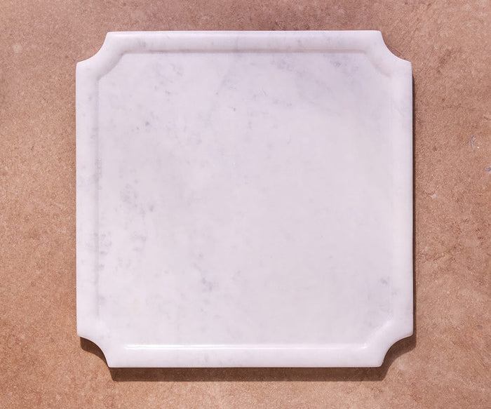 The Vivaen Framed Square Board, crafted from white marble with beveled edges and notched corners, rests on a light brown textured surface—an elegant choice for serving.