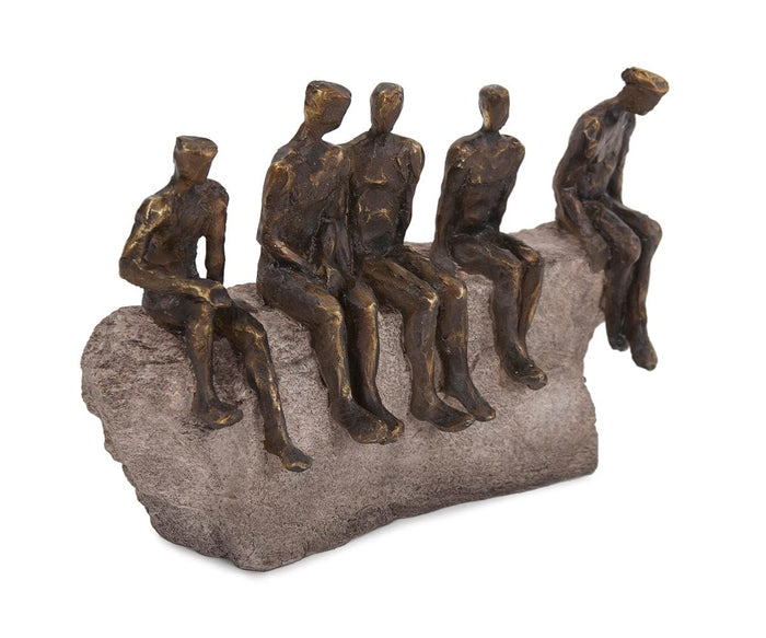 Five faceless, minimalist bronze figurines with a bronze finish are seated closely together on a rough stone-like bench, their casual interaction capturing the abstract style of the Waiting Figure Statue.