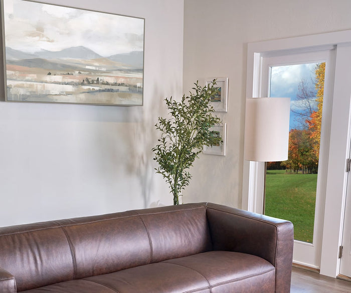 A brown leather couch sits near a white wall adorned with serene decor, including the Whisper Valley Wall Art landscape painting. A lamp and potted plant are by a glass door, which reveals a grassy yard and colorful fall trees outside.
