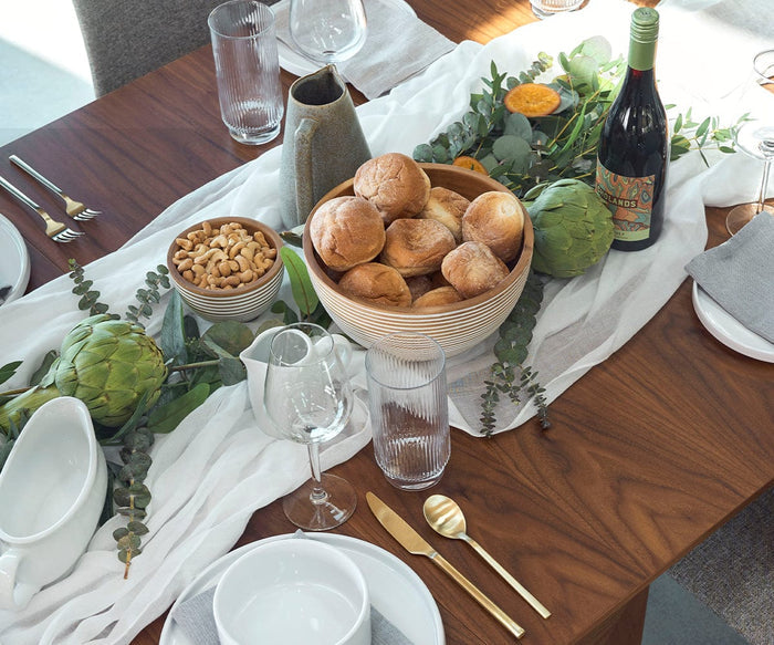 A wooden table set for a meal features handcrafted Winifred Bowls made from sustainably sourced mango wood, filled with bread rolls and cashews, alongside artichokes, a wine bottle, glasses, plates, gold flatware, and decorative greenery—all beautifully arranged for an inviting dining experience.