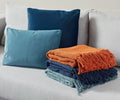 Two blue pillows rest on a light grey couch, adding a touch of mid-century color to the space. In front, three folded, fringed Woven Fringe Throw blankets in rust are stacked neatly on the sofa cushion, highlighting contemporary texture and style.