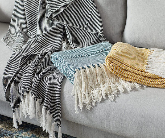 A black Woven Twill Throw with white stripes is folded on a light gray couch. This decorative cotton throw features a woven twill design and is finished with white fringe along the edges.
